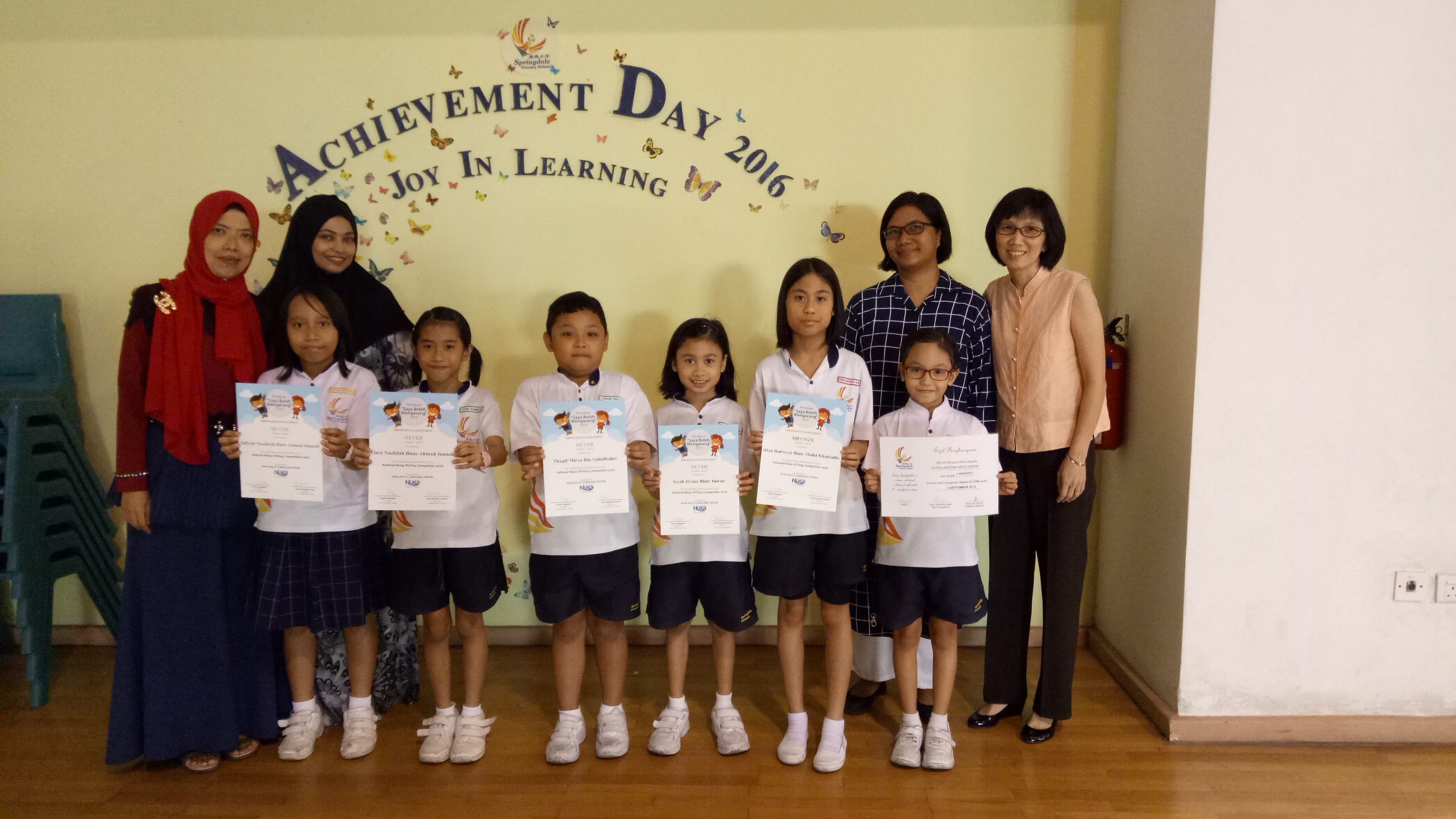 Six children in school uniforms hold certificates in front of a wall reading Achievement Day 2016 Joy In Learning.
